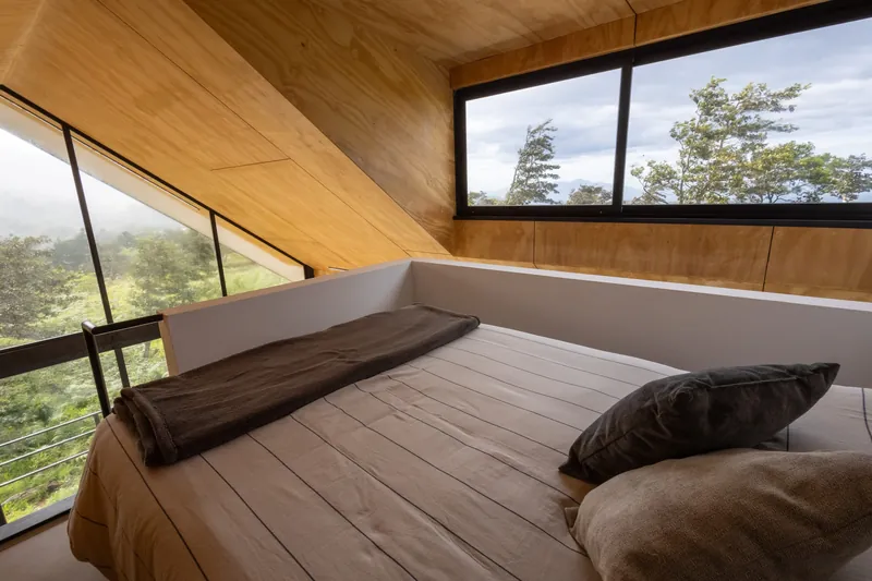 Cama queen en el mezzanine de madera, con una ventana larga hacia los árboles
